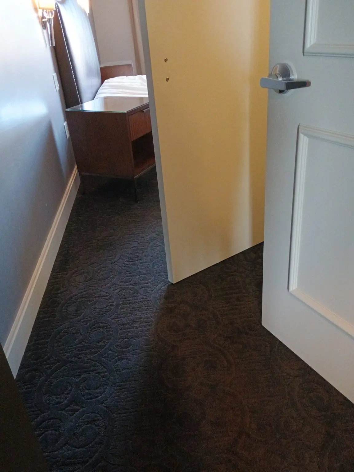 Open door into a hotel bedroom; a bed and wooden nightstand are visible in the room beyond the doorway.