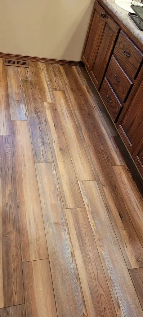 Wide view of a warm wooden kitchen floor with brown cabinets along the right wall and a floor vent near the baseboard.